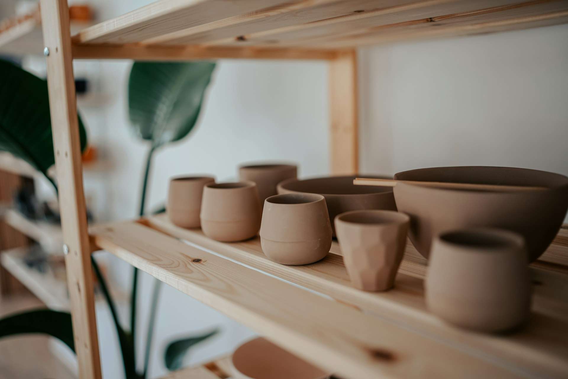 Unglazed ceramic cups, bowls and vessels drying on a wooden studio shelf.