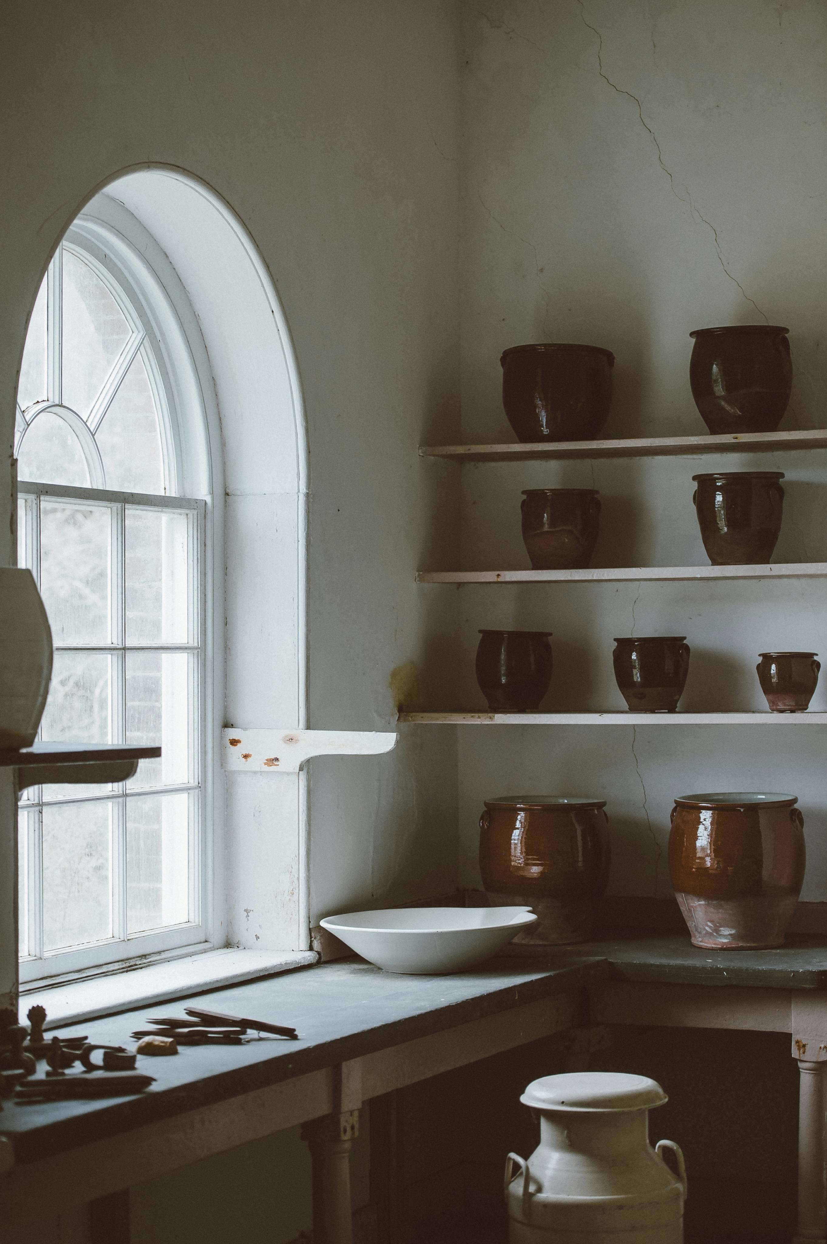Glazed brown ceramic vessels displayed on wooden shelves beside a large arched studio window.